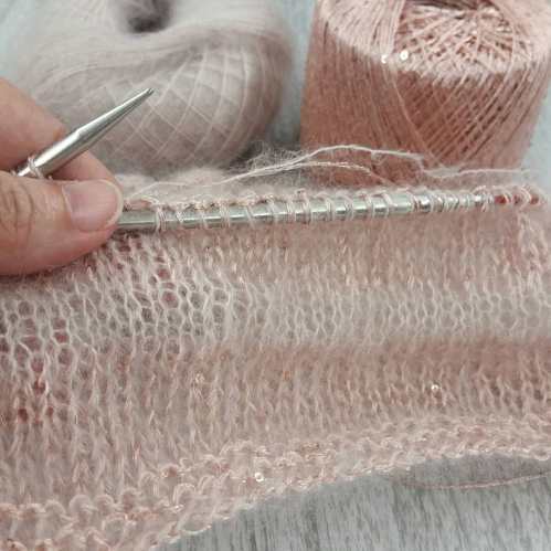 A close-up of a hand holding knitting needles with soft pink yarn and a partially knitted piece in the foreground.