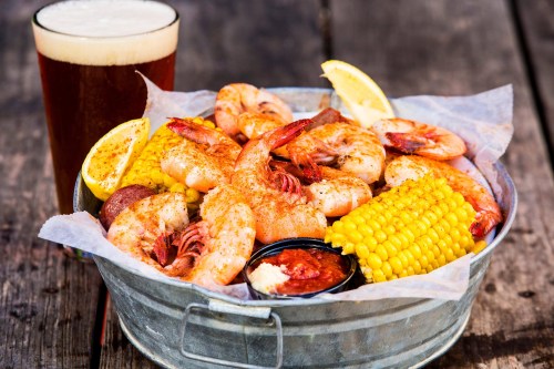 A metal bucket filled with seasoned shrimp, corn, and a side of sauce, accompanied by a glass of beer.