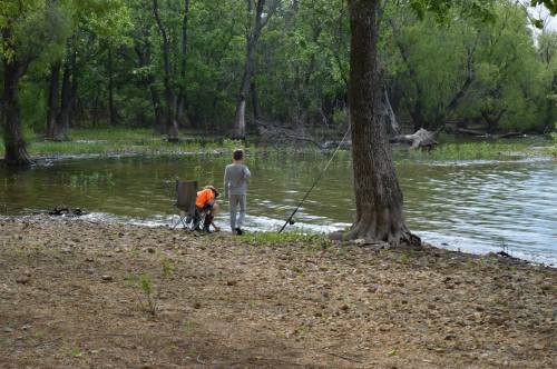 Two children fishing by a calm river surrounded by lush greenery and trees.