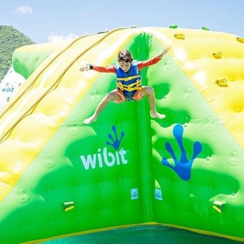 A child in a life jacket jumps off a colorful inflatable water obstacle, surrounded by blue skies and water.