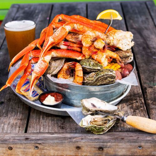 A metal platter filled with various seafood, including crab legs, shrimp, oysters, and a lemon wedge, on a wooden table.
