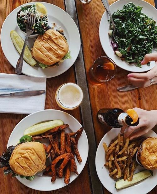 A table with two plates of burgers, sides of fries, a salad, and drinks, all on a wooden surface.