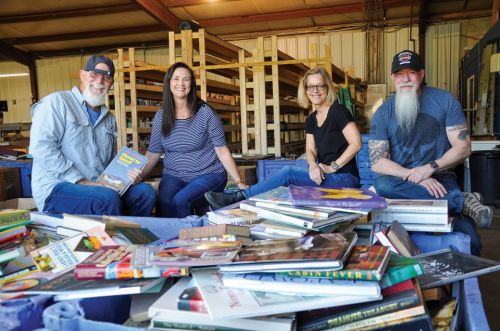 Four people sit around a table filled with books in a warehouse, smiling and engaging with each other.