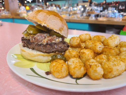 A close-up of a burger with pickles on a plate, accompanied by crispy tater tots, set on a pink table.