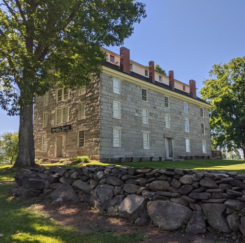Historic stone building with large windows, surrounded by trees and a stone wall, under a clear blue sky.