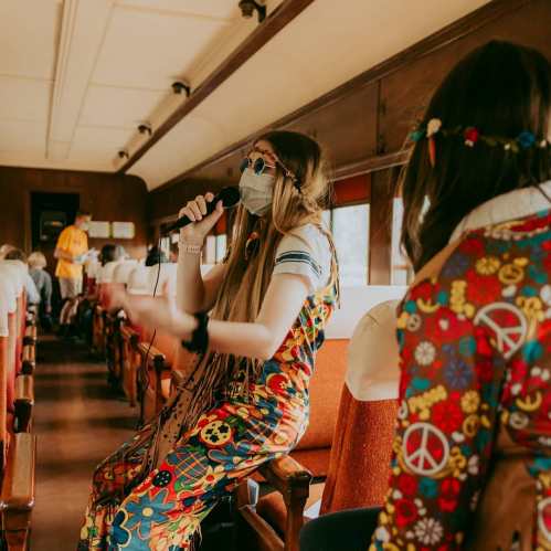 A performer in a colorful outfit sings into a microphone on a vintage train, while passengers enjoy the show.