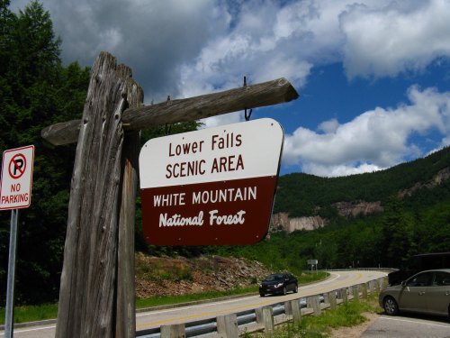 Sign for Lower Falls Scenic Area in White Mountain National Forest, with a cloudy sky and mountains in the background.