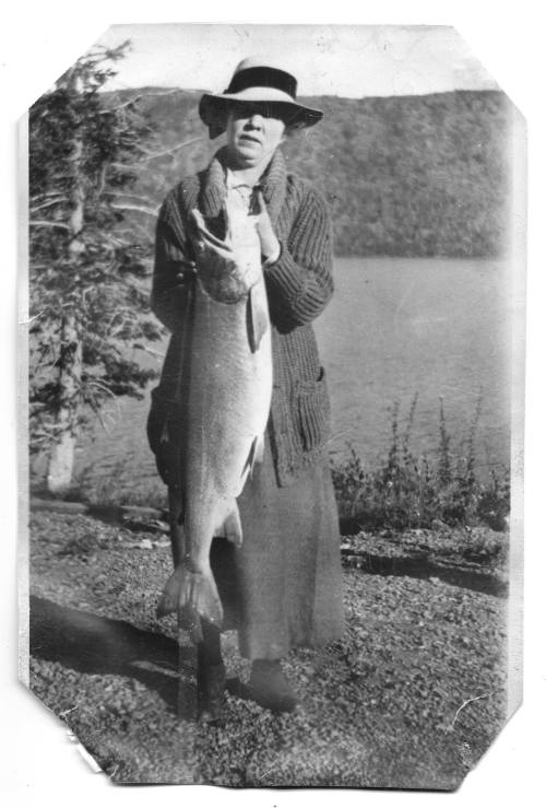 A woman in a hat holds a large fish by a lake, surrounded by trees, in a vintage black-and-white photo.