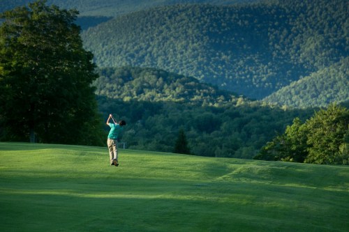 A golfer swings on a lush green course with mountains in the background under a clear blue sky.