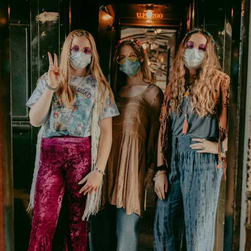 Three women in colorful outfits and masks pose together in a vintage train car, showcasing a retro style.