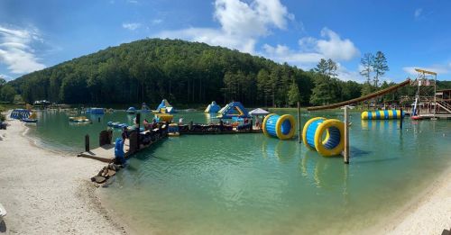 A scenic lake with inflatable water attractions, surrounded by trees and a sandy beach under a blue sky.
