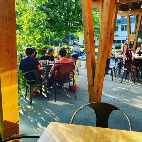 A group of people sitting at an outdoor café, surrounded by greenery and wooden structures, enjoying their time together.