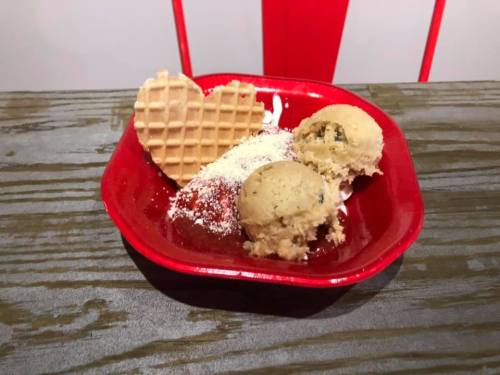 A red dish holds two scoops of cookie dough ice cream, a strawberry, and a heart-shaped waffle.