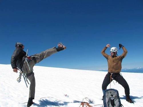 Two climbers on a snowy peak, one kicking playfully while the other raises arms in excitement against a clear blue sky.
