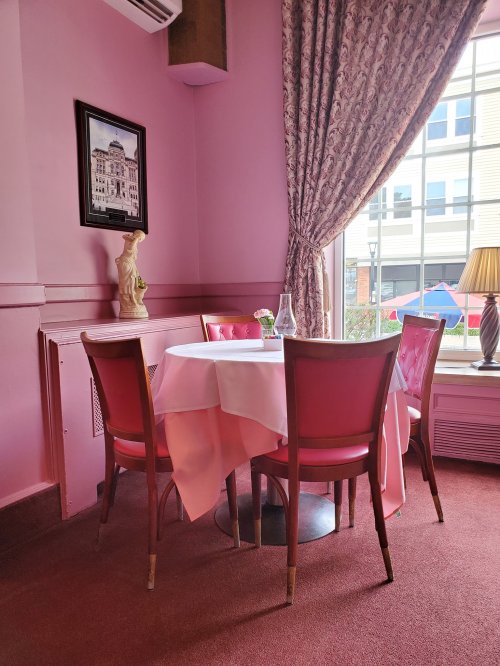 A cozy pink dining area with a round table, pink chairs, and a large window letting in natural light.