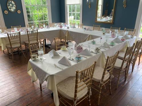 A beautifully set dining room with tables draped in white cloth, elegant chairs, and tea sets ready for a gathering.