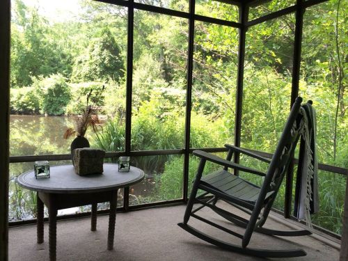 A cozy screened porch with a rocking chair and table, overlooking a lush green landscape and a tranquil pond.