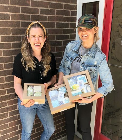 Two smiling women hold boxes of treats outside a brick building, wearing casual outfits and accessories.