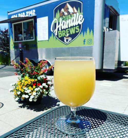 A glass of yellow drink sits on a table in front of a colorful food truck named "Handle Brews." Flowers are in the background.