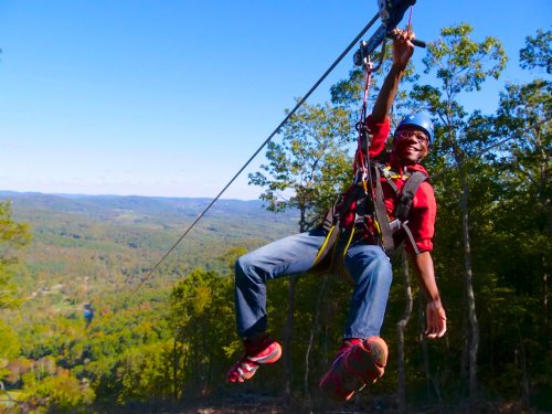 A person zip-lining through a scenic landscape with trees and mountains in the background, smiling and enjoying the adventure.