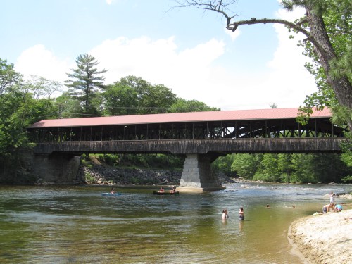 A covered bridge spans a river, with people swimming and kayaking nearby, surrounded by lush greenery and blue skies.