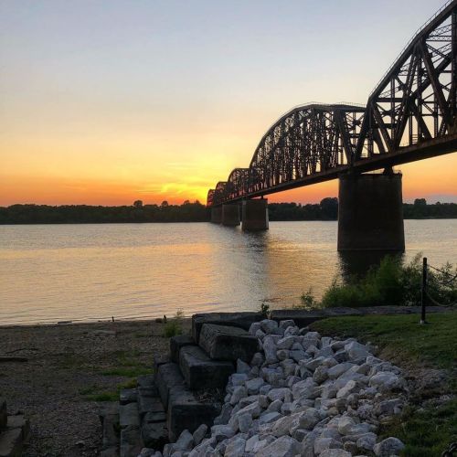 A sunset view of a bridge over a calm river, with rocks in the foreground and trees lining the shore.