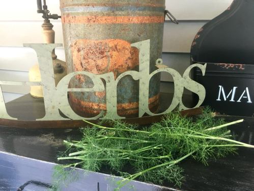 A decorative "Herbs" sign with fresh dill in front of a rustic container and a black box labeled "MA."