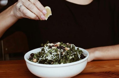 A person squeezing lemon over a bowl of kale salad topped with nuts and cheese.