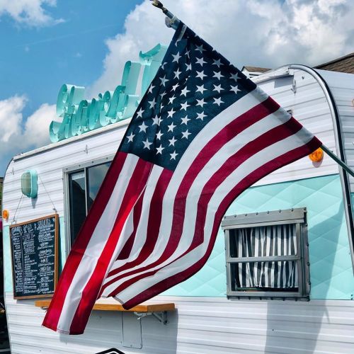 A vintage food truck with a turquoise sign and an American flag waving in front against a cloudy sky.