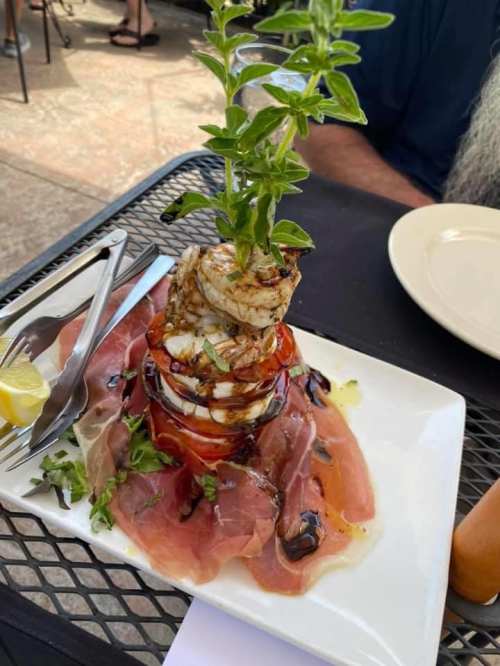 A gourmet dish featuring grilled shrimp atop layers of tomato, prosciutto, and greens, drizzled with balsamic glaze.