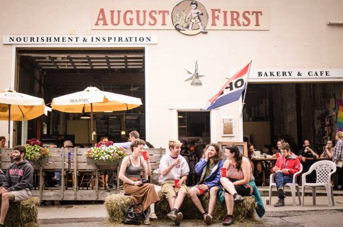 A lively café scene with people sitting on hay bales outside August First, enjoying food and conversation.