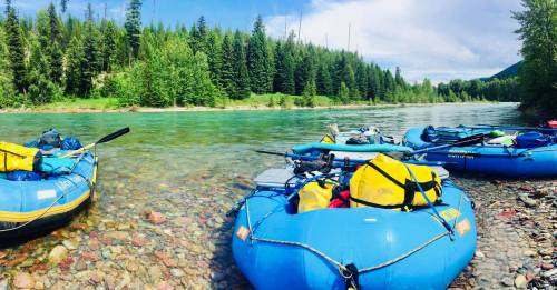 Rafts with gear are moored by a clear river, surrounded by lush green trees and a bright blue sky.