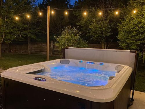 A lit hot tub in a backyard at dusk, surrounded by trees and string lights, creating a cozy atmosphere.