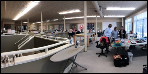 A spacious craft room with several tables, people working on projects, and large windows providing natural light.