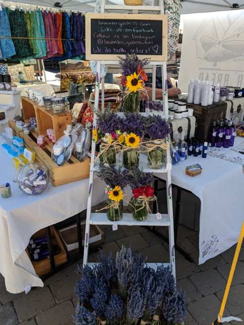 A display of lavender and sunflower arrangements on a ladder at a market, with various products in the background.