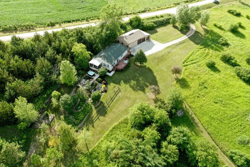 Aerial view of a house surrounded by greenery, with a driveway and garden, near a rural road.