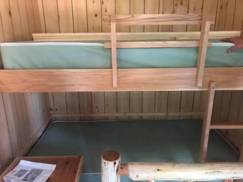 A wooden bunk bed with a green mattress on the top and a lower bed frame, set against a wooden wall.