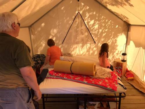 Two adults and a child inside a tent, arranging bedding on two beds with colorful blankets and a lantern nearby.