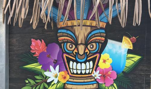 Colorful tiki mask surrounded by tropical flowers and a cocktail, set against a wooden background with a thatched roof.