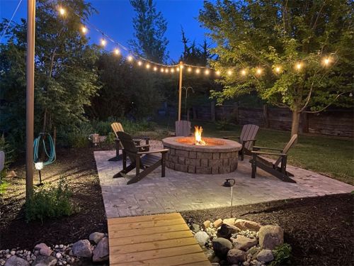 Cozy outdoor fire pit area with string lights, surrounded by chairs and greenery, set in a tranquil evening scene.