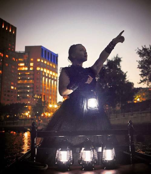 A woman in a black dress stands on a boat, pointing upward with lanterns glowing around her at dusk.
