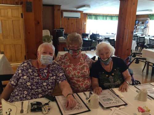Three elderly women wearing masks sit together at a dining table, smiling and enjoying their time in a cozy restaurant.