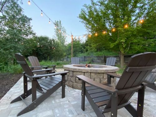 A cozy outdoor fire pit area with wooden chairs, surrounded by greenery and string lights overhead.