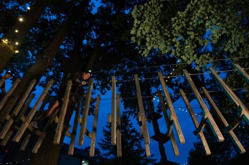 A person navigates a wooden rope course among trees at dusk, with string lights illuminating the scene.