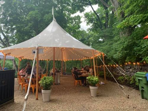 A large white tent with string lights, surrounded by trees, hosting people dining at tables on a gravel surface.