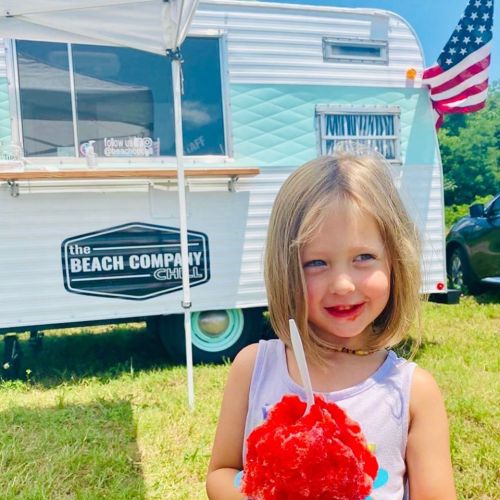 A young girl with a short haircut smiles while holding a red snow cone in front of a vintage food truck.