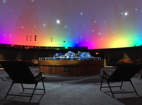 A planetarium interior with colorful lights and a central display, featuring two empty chairs in the foreground.
