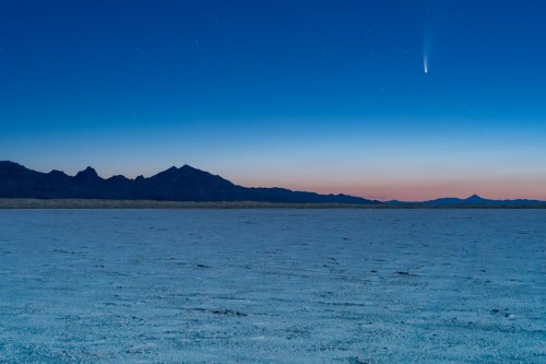 A comet streaks across a twilight sky above a vast, salt flat and distant mountains.