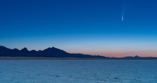 A comet streaks across a twilight sky above a vast, flat landscape and distant mountains.