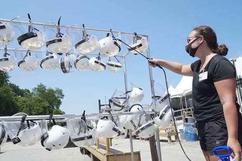A person wearing a mask cleans safety helmets on a rack outdoors under a clear blue sky.